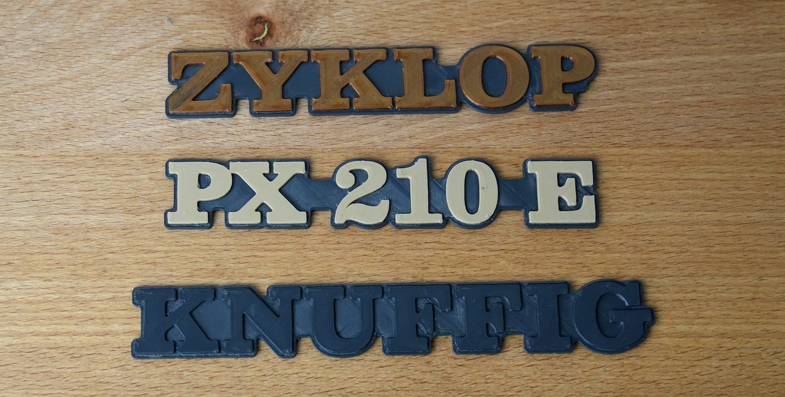 Vier fertig gedruckte Schriftzüge auf einem Holztisch: ZYKLOP in Kupfer, PX 210 E in Creme, KNUFFIG in Grau und CELENTANO in Dunkelrot, jeweils auf grauem Träger